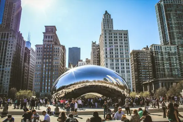 Crowd of people gathered around the reflective Cloud Gate sculpture, also known as The Bean, in Millennium Park, Chicago, with skyscrapers and clear blue sky in the background.
