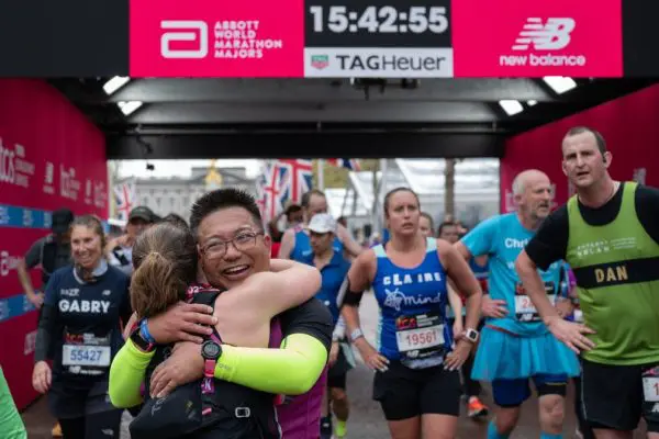 A group of runners crossing the finish line of the TCS London Marathon. Two participants are hugging each other.